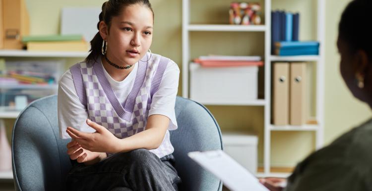 A young person sat in a chair talking to a person holding a piece of paper opposite them