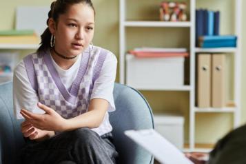 A young person sat in a chair talking to a person holding a piece of paper opposite them 